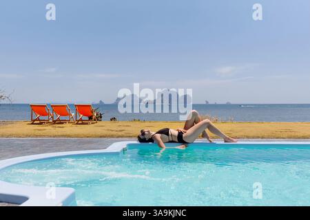 Eine Person genießt Freizeit, indem sie im Liegestuhl in einem Pool schwimmt. Meereswellen schlagen sanft gegen das Ufer, und entfernte Inseln sind unter A zu sehen Stockfoto