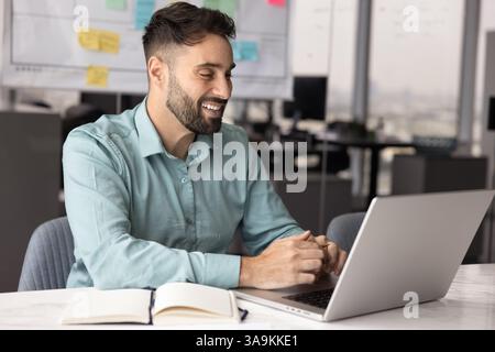 Positiver hispanischer Geschäftsmann, der Laptop für die Kommunikation am Arbeitsplatz benutzt Stockfoto