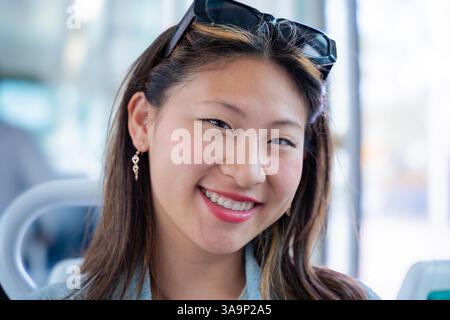 Lächelnde Chinesin, die in die Kamera schaut, während sie auf dem Sitz für öffentliche Verkehrsmittel sitzt Stockfoto