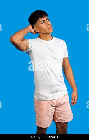 Nachdenklicher junger latino-Mann in weißem T-Shirt und rosa Shorts, der sein Haar berührt, während er auf blauem Hintergrund aufblickt Stockfoto