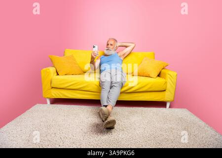 Seniorenmann, der sich auf einem gelben Sofa in einem leuchtenden rosafarbenen Zimmer entspannt und einen zwanglosen und stilvollen Lifestyle-Moment festgehalten hat Stockfoto