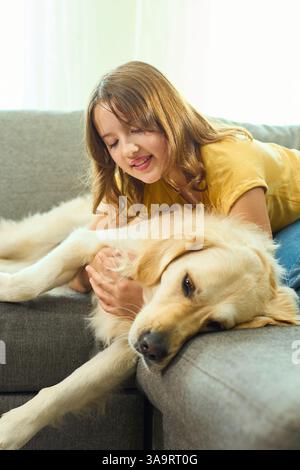 Ein fröhliches Teenager-Mädchen kuschelt ihren goldenen Retriever auf einer weichen Couch in einem hellen Wohnzimmer. Sonnenlicht strömt herein und schafft eine warme Atmosphäre Stockfoto