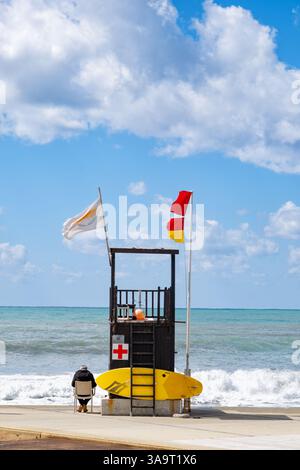 Pathos, Zypern. Ein Rettungsschwimmer bemannt einen Rettungsschwimmer-Turm an einem beliebten Strand. Starke Winde und raue See bedeuten, dass rote Warnflaggen fliegen, um die Wassernutzer zu warnen Stockfoto