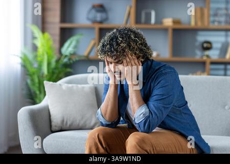 Gestresster Mann mit lockigem Haar, der auf einer Couch sitzt und in einer modernen Einrichtung schwere Kopfschmerzen hat. Er ist von negativen Emotionen überwältigt. Stockfoto