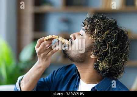 Junger zufriedener Mann zu Hause, der Pizza isst, während er auf dem Sofa im Wohnzimmer der Wohnung sitzt. Stockfoto