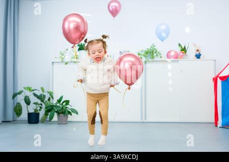 Niedliches 3-jähriges Mädchen mit Down-Syndrom feiert ihren Geburtstag in einem mit Ballons dekorierten Spielzimmer. Kindergeburtstag. Stockfoto