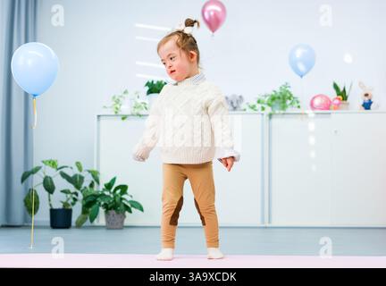 Niedliches 3-jähriges Mädchen mit Down-Syndrom feiert ihren Geburtstag in einem mit Ballons dekorierten Spielzimmer. Kindergeburtstag. Stockfoto