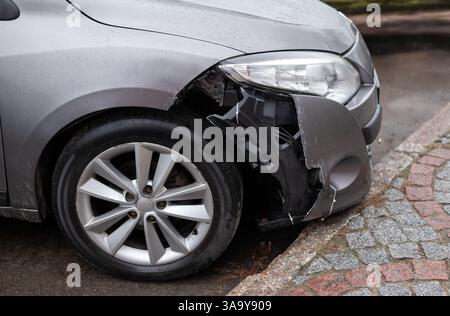 Vorderseite eines silbernen Autos mit beschädigtem Stoßfänger und freiliegender Innenstruktur nach einem Unfall, parkt in der Nähe einer Bordsteinkante auf einer nassen Straße Stockfoto
