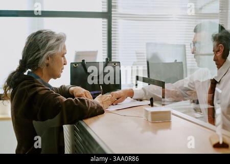 Ältere Frau diskutiert und unterzeichnet Dokumente an modernen Bürotheken, während junger Mann hinter Glaswand, Büroausstattung und Taschen im Hintergrund hilft Stockfoto