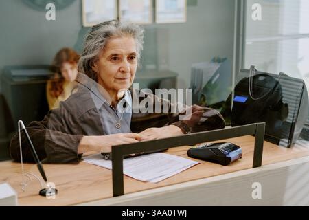 Ältere Frau, die mit Papieren am Schreibtisch sitzt, an der Arbeit beteiligt ist, verschwommener Hintergrund mit einer anderen Person und Büroausstattung, was auf ein professionelles Umfeld hinweist Stockfoto