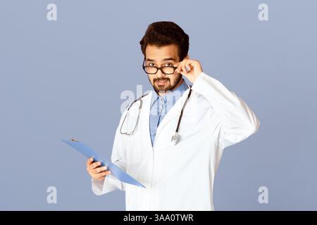 Junge indische Arzt in Uniform und Brille hält Zwischenablage an Violetter Hintergrund Stockfoto