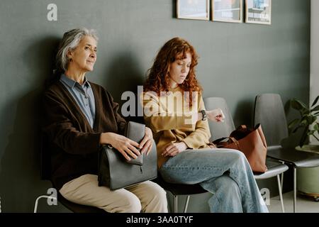 Zwei Frauen sitzen im Wartebereich des Büros, eine mit Mappe und Bürokleidung, die andere mit legeren Klamotten Stockfoto