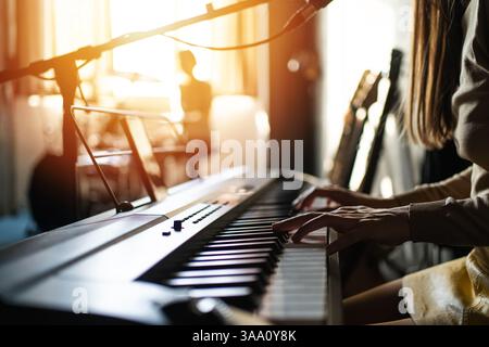 Frau, die digitales Klavier im sonnendurchfluteten Musikstudio spielt, umgeben von Gitarren und Aufnahmegeräten, Konzept der Kreativität, Musikproduktion, Lifestyle und Künstlerinnen. Stockfoto