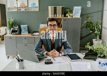 Porträt eines Geschäftsmannes, der eine Brille trägt, lächelt und in einer modernen Büroumgebung am Schreibtisch sitzt, mit verschiedenen Büromaterialien, Gadgets und Pflanzen, die ihn umgeben Stockfoto