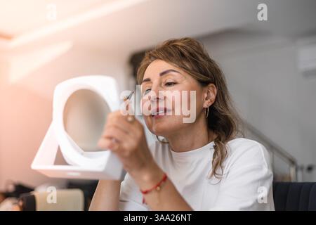 Eine Frau sitzt in einem stilvollen Raum, verwendet einen kompakten Spiegel, eine Pinzette und entfernt Augenbrauen. Natürliches Licht beleuchtet ihren fokussierten Ausdruck als s Stockfoto