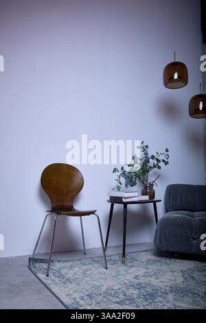 Minimalistisches Wohnzimmer mit grauem Sofa, elegantem Stuhl und einem kleinen Tisch mit Pflanzen. Drei Hängeleuchten hängen darüber und sorgen für ein gemütliches und modernes Ambiente Stockfoto