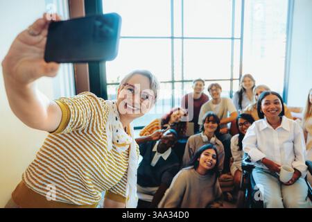 Eine lebhafte Gruppe von Schülern posiert fröhlich mit ihrem Mentor und macht ein fröhliches Selfie in einem hellen Klassenzimmer. Stockfoto