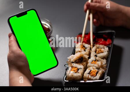 Frau, die zu Hause Sushi-Brötchen isst. Smartphone mit grünem Bildschirm mit Platz für Text. Smartphone in der Hand mit Serviertisch in b Stockfoto