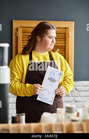 In einem lebhaften Café präsentiert eine junge Frau mit Down-Syndrom stolz das Menü, während sie lächelt. Stockfoto