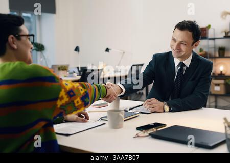 Geschäftsmann schüttelt dem Mitarbeiter die Hand, während er während des Meetings am Tisch sitzt Stockfoto