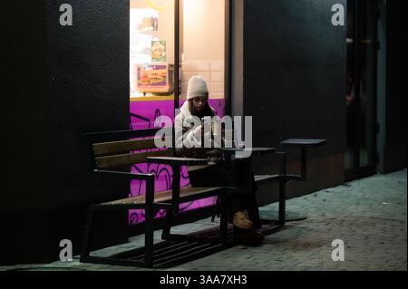 Eine junge Frau sitzt nachts leise auf einer Bank, in ihr Handy eingesogen. Das leuchtende Licht eines nahe gelegenen Fensters erzeugt ein warmes Licht, das die Dunkelheit kontrastiert Stockfoto