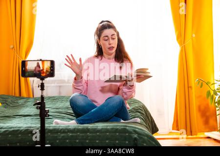 Junge kaukasische Frau filmt sich zu Hause, während sie auf einem Bett sitzt und digitale Inhalte für ihre Social-Media-Präsenz erstellt. Mitten im Shot. Stockfoto