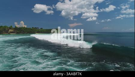 Atemberaubender Blick aus der Vogelperspektive mit türkisfarbenen Meereswellen, die am Dreamland Beach in Bali brechen, eingerahmt von üppigen grünen Klippen und weißen Wolken, schaffen ein tropisches Paradies Stockfoto