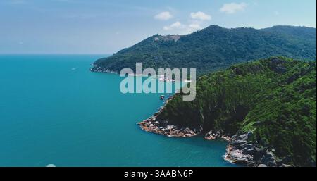 Türkisfarbenes Wasser streichelt sanft die felsigen Küsten einer üppigen tropischen Insel, mit einem kleinen Boot, das durch das ruhige Meer und einen grünen Berg als atemberaubende Kulisse navigiert Stockfoto