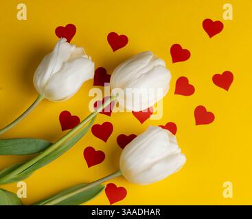 Drei frische und schöne weiße Tulpen auf gelbem Papier mit roten Herzen, roten Herzen und weißen Tulpenblüten auf gelbem Papier, Nahaufnahme Stockfoto