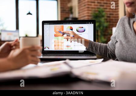 Weibliche Beraterin, die während des Gesprächs mit der Kollegin auf den Laptop zeigt, um Geschäftsanalysen anzuzeigen. Selektiver Fokus auf ein digitales Gerät am Bürotisch, das Unternehmensdaten-Diagramme anzeigt. Stockfoto