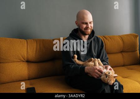 Fröhlicher junger erwachsener Mann mit Bart, der legere Kleidung trägt, liebevoll mit Ingwerkatze spielt, einen ruhigen Moment zu Hause genießt, auf dem Sofa sitzt. Stockfoto