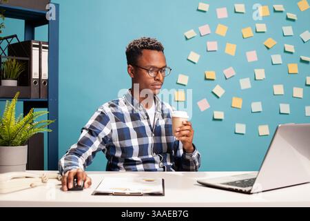 afroamerikanischer Startup-Mitarbeiter konzentrierte sich auf den Laptop-Bildschirm und analysierte geschäftliche Kennzahlen an seinem Schreibtisch. Marketingspezialist hält eine Maus und eine Kaffeetasse, die online surft und Forschungsdaten überprüft. Stockfoto