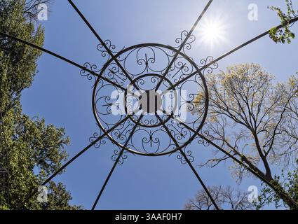 Blick nach oben durch ein dekoratives eisernes Pavillondach in Richtung einer hellen Sonne in einem wolkenlosen Himmel, der im Frühling von Bäumen umrahmt wird. Stockfoto