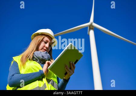 Ingenieurin, die ein digitales Tablet neben Windkraftanlagen in einer Farm für erneuerbare Energien verwendet Stockfoto