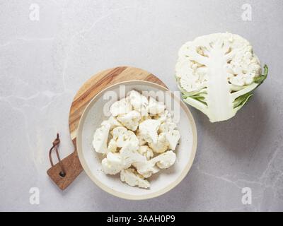 Roher frischer Blumenkohl, der auf der Arbeitsplatte neben einer Schüssel auf einem Holzschneidebrett mit in Röschen geschnittenen Stückchen in zwei Hälften geschnitten wird Stockfoto