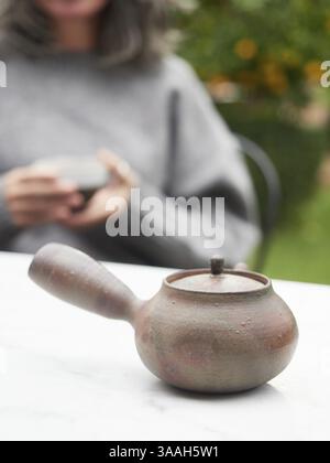 Traditionelle japanische Teekanne mit seitlichem Griff, genannt Kyusu. Unfokussierter Hintergrund unerkennbare Frau, die den Becher mit den Händen hält Stockfoto