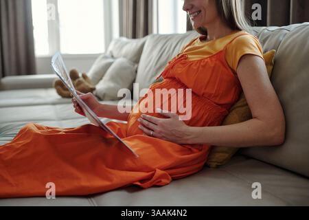 Glückliche schwangere Frau, die das Magazin auf der Couch liest Stockfoto