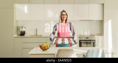 Eine junge Frau mit einer Schürze, die ein Tablett mit gebackenen Keksen in der Küche hält Stockfoto