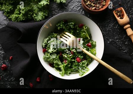 Schüssel mit leckerem Grünkohl-Salat mit getrockneten Preiselbeeren, Gabel und Pfefferkörnern auf dunklem Hintergrund Stockfoto