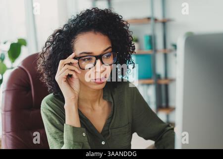 Junge Frau, die an einem Schreibtisch arbeitet, während sie ihre Brille anpasst und sich auf eine Aufgabe konzentriert Stockfoto