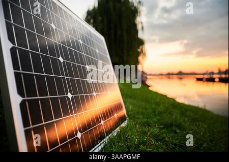 Sonnenkollektor, beleuchtet von der untergehenden Sonne, steht am grasbewachsenen Seeufer, mit malerischem Sonnenuntergang und Bäumen im Hintergrund. Fusion erneuerbarer Energietechnologien mit ruhiger Naturlandschaft. Stockfoto