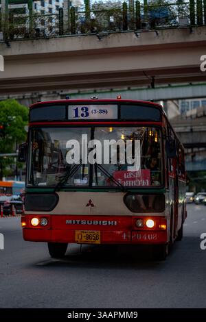 Dieses nostalgische Bild in der Abenddämmerung zeigt einen alten Mitsubishi Bus auf der Ratchadamri Road in Bangkok, Thailand, der durch die geschäftigen Straßen der Stadt fährt. Rot und Creme- Stockfoto