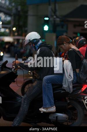 Dieses lebhafte Bild in der Abenddämmerung fängt zwei Personen auf einem Moped ein, die beide in ihre Handys vertieft sind, an der Rama 1 Road in Bangkok, Thailand. Die Szene, inszeniert auf Stockfoto