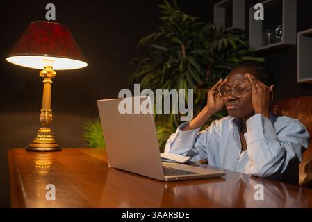 Professionelle afroamerikanische Geschäftsfrau leidet unter arbeitsbedingtem Stress, drückt in schwach beleuchtetem Büro Schläfen in der Nähe des Laptops und leidet während des späten Nachtprojekts unter intensiven Kopfschmerzen Stockfoto