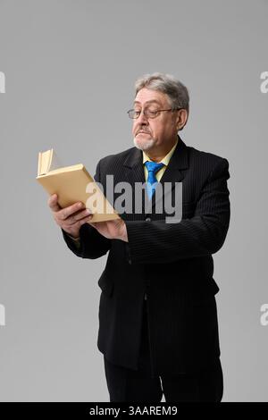 Senior Mann, Professor im Anzug, der ein Buch mit ernstem Ausdruck auf grauem Hintergrund hält. Stockfoto