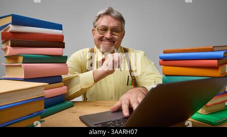 Fröhlicher älterer Mann in Fliege, der am Schreibtisch sitzt, lächelt und auf dem Laptop zwischen den Büchern schreibt. C Stockfoto