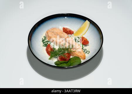 Gegrilltes Lachsfilet, serviert mit Spinat-Cremesauce, Kirschtomaten, Dill und Zitronenkeil in einer Keramikschale. Stockfoto