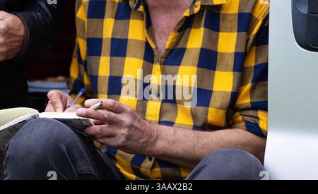Nahaufnahme eines Mannes in einem gelben karierten Hemd, der einen Stift und eine Zigarette hält, im Wohnwagen sitzt und in Notizbuch schreibt. Stockfoto