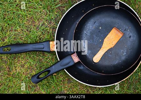 Blick von oben auf zwei schwarze Bratpfannen mit Holzspachtel auf grünem Gras, symbolisiert Campingstil, Outdoor-Küche, Minimalismus und rustikale Lebensweise Stockfoto