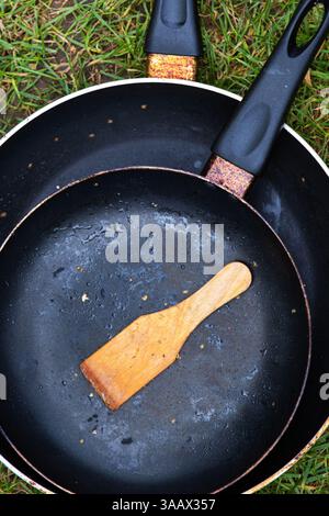 Blick von oben auf zwei schwarze Bratpfannen mit Holzspachtel auf grünem Gras, symbolisiert Campingstil, Outdoor-Küche, Minimalismus und rustikale Lebensweise Stockfoto